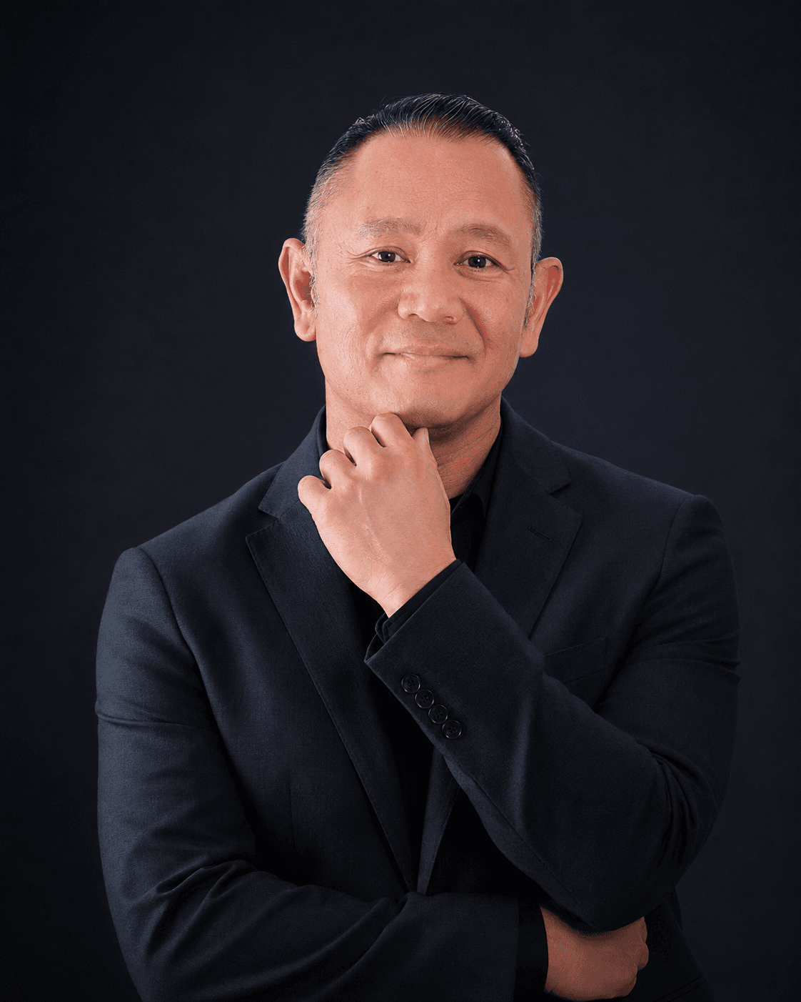 Smiling Asian man in a navy suit with hand on chin against black background.